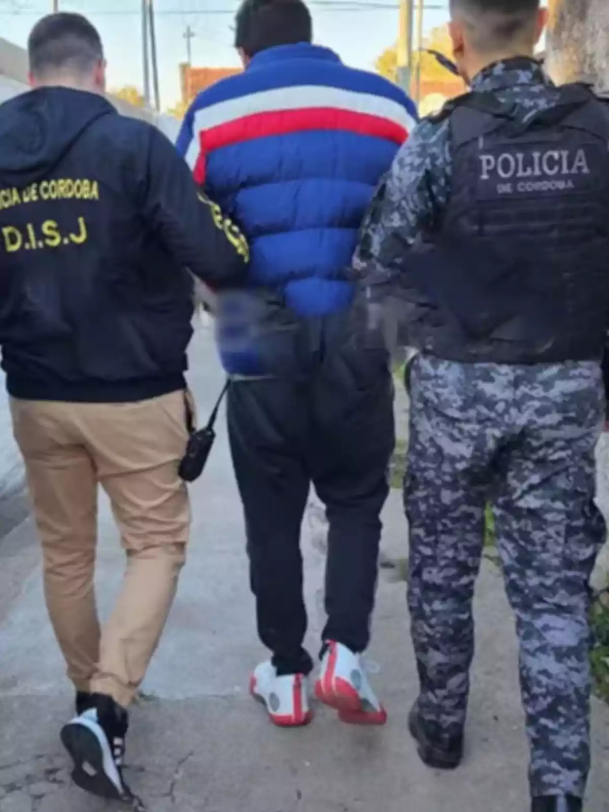 Tres personas caminando por una vereda, dos de ellas con uniformes policiales escoltan a un hombre con campera azul