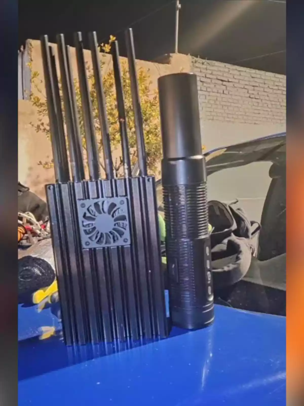 Dispositivo inhibidor de señal con varias antenas junto a una linterna negra sobre una superficie azul Dispositivo inhibidor de señal con varias antenas junto a una linterna negra sobre una superficie azul