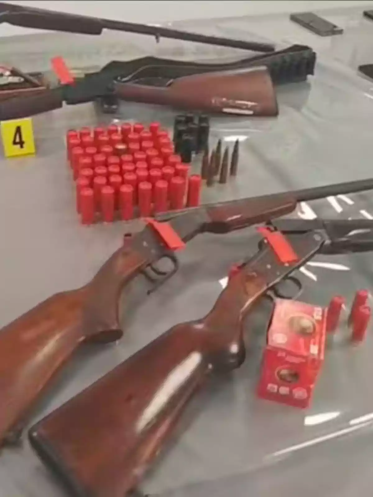 Three shotguns made of wood and metal next to red cartridges and bullets on a glass table, with a yellow tag labeled number 4 Three shotguns made of wood and metal next to red cartridges and bullets on a glass table, with a yellow tag labeled number 4