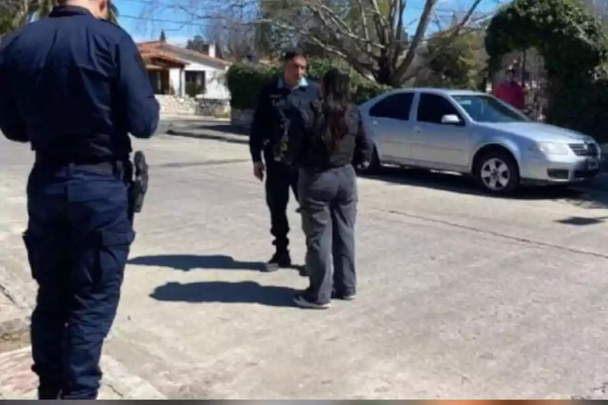 Two police officers and a woman are talking on a residential street next to a parked silver car.