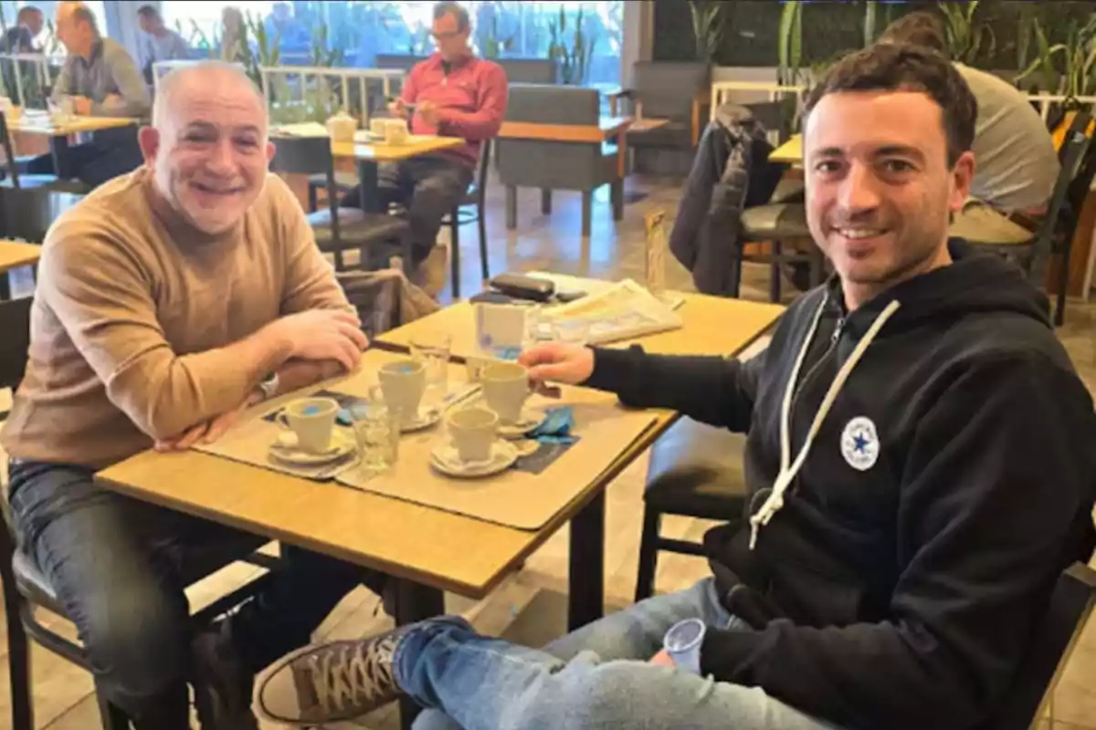Two men smile while enjoying coffee at a café, sitting at a table with cups and glasses, with other people in the background. Two men smile while enjoying coffee at a café, sitting at a table with cups and glasses, with other people in the background.