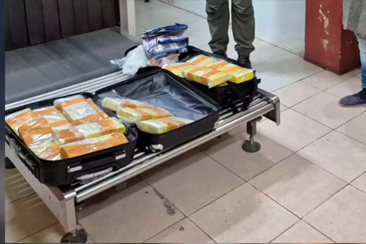 Maletas abiertas sobre una banda de equipaje con varios paquetes envueltos en plástico y cinta adhesiva en su interior, junto a personas de pie. Maletas abiertas sobre una banda de equipaje con varios paquetes envueltos en plástico y cinta adhesiva en su interior, junto a personas de pie.