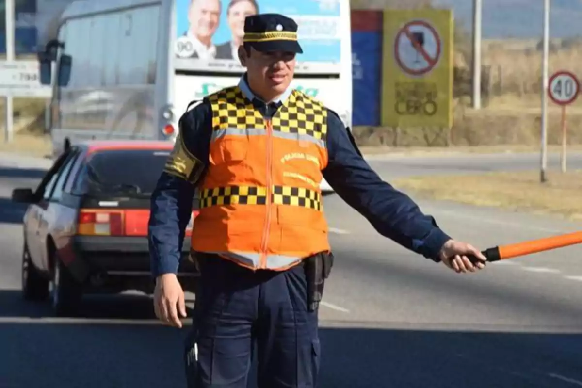 Oficial de tránsito con chaleco naranja y gorra dirigiendo el tráfico en una carretera mientras sostiene una vara señalizadora