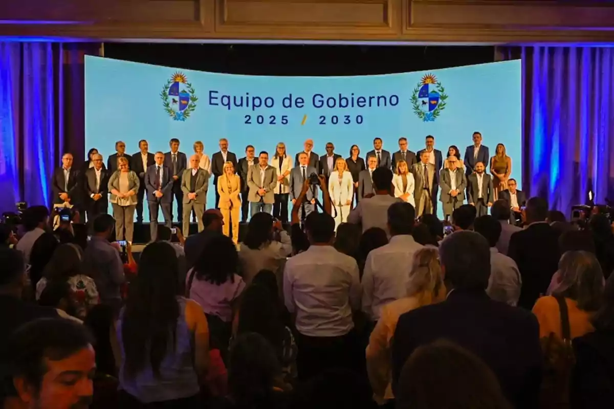 A large group of people posing on a stage in front of an audience with a banner in the background that reads Government Team 2025 2030 and the national emblems on the sides A large group of people posing on a stage in front of an audience with a banner in the background that reads Government Team 2025 2030 and the national emblems on the sides