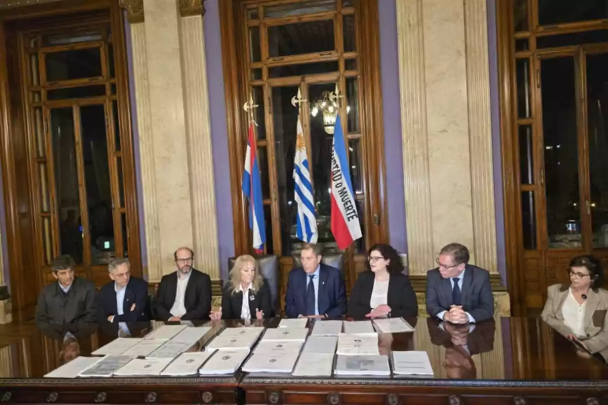 Eight people seated at a wooden table with documents in front of them in an elegant room with large windows and flags in the background