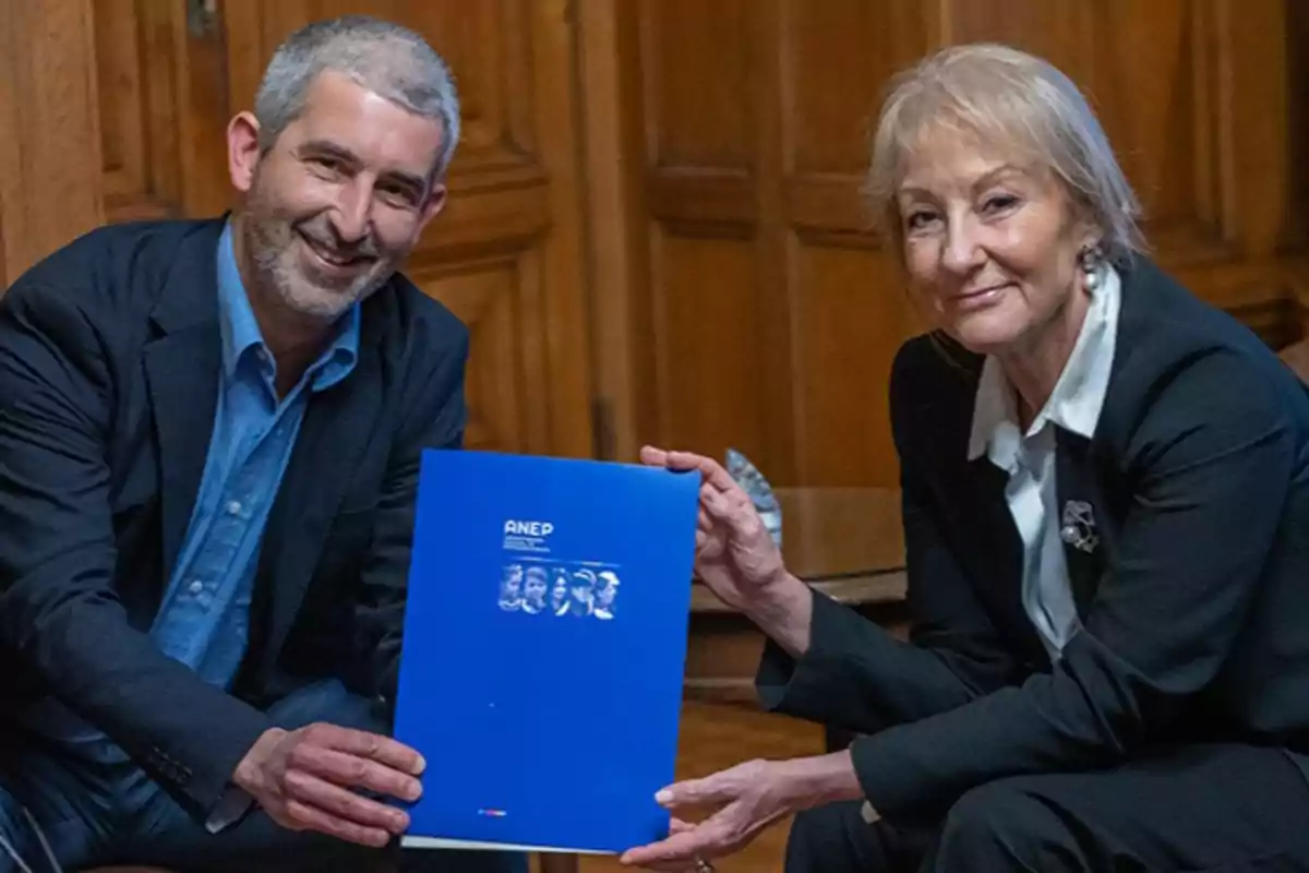 Two people smile while holding a blue ANEP folder together in a wooden office.