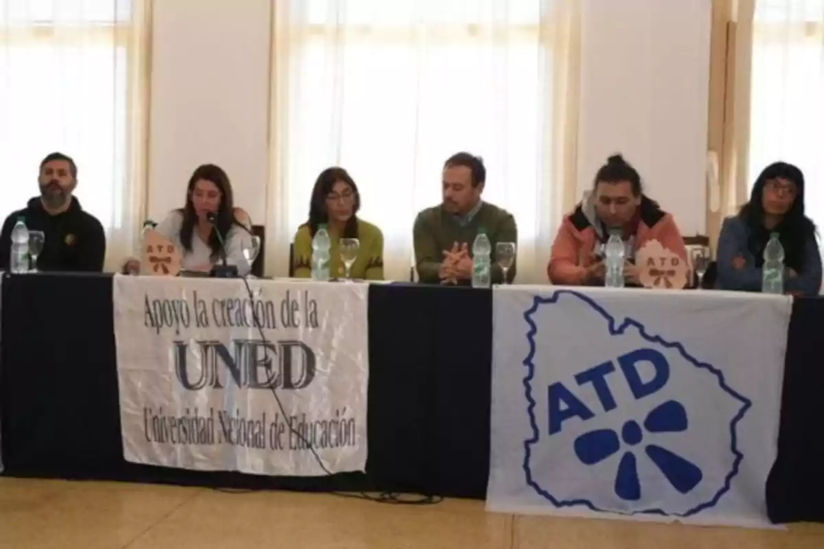 Seis personas sentadas en una mesa larga durante una conferencia, con botellas de agua y copas, y dos pancartas que apoyan la creación de la Universidad Nacional de Educación y la organización ATD