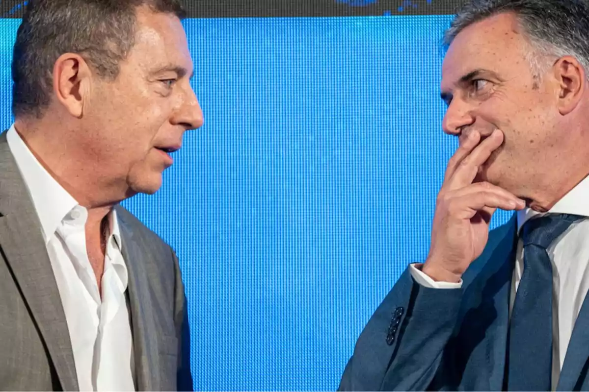 Two men in suits are talking in front of a blue screen.