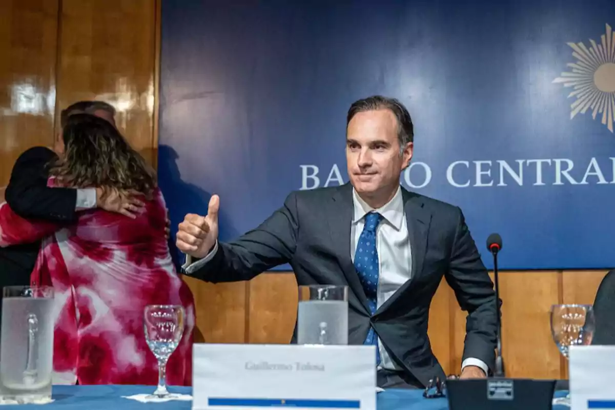 Hombre de traje sentado en una mesa con un cartel que dice Guillermo Tolosa levantando el pulgar mientras al fondo dos personas se abrazan frente a un cartel azul que dice Banco Central