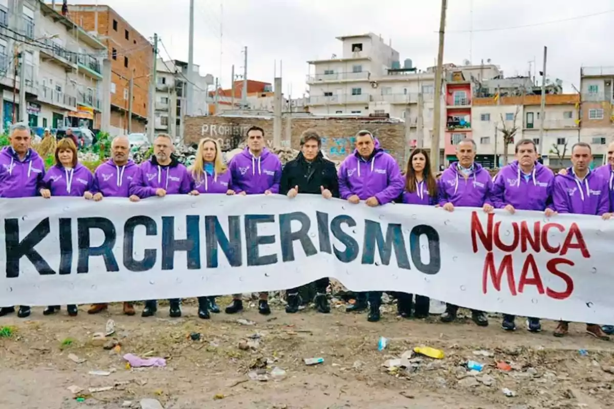 Un grupo de personas con camperas violetas sostiene una pancarta que dice Kirchnerismo nunca más en un barrio urbano con edificios de fondo. Un grupo de personas con camperas violetas sostiene una pancarta que dice Kirchnerismo nunca más en un barrio urbano con edificios de fondo.