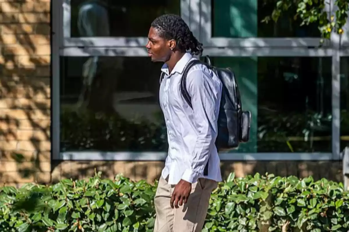 Young man walking outdoors wearing a white jersey and a black backpack in front of a building with large windows and green bushes