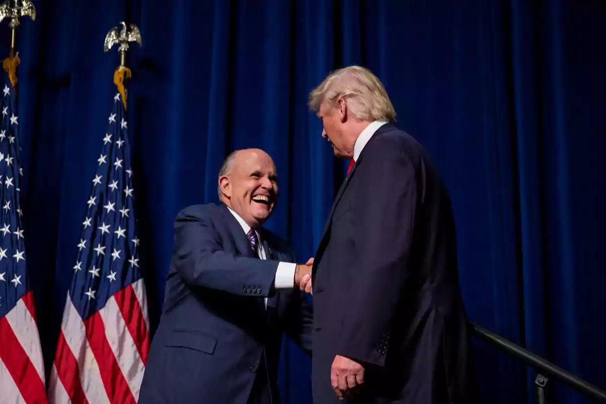 Dos hombres de traje se dan la mano sonrientes frente a un fondo azul con banderas de Estados Unidos