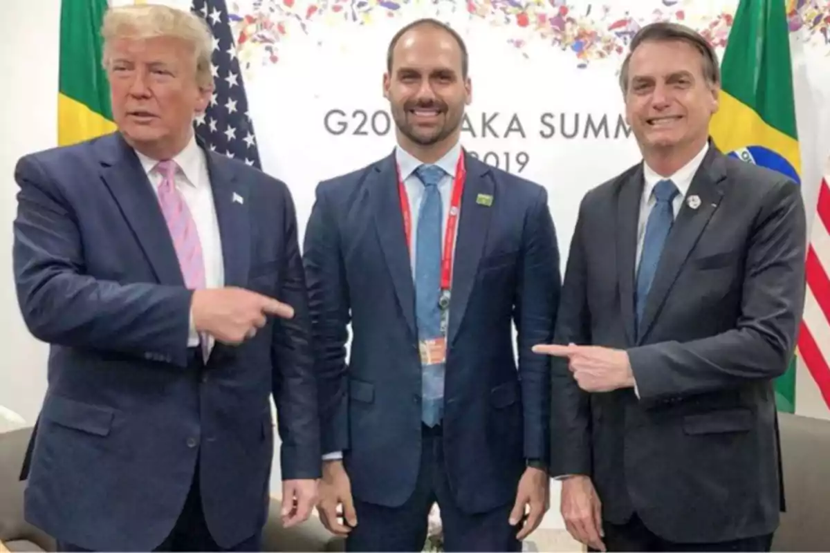 Tres hombres con traje posan para una foto en un evento internacional con banderas de fondo y dos de ellos señalan al hombre en el centro