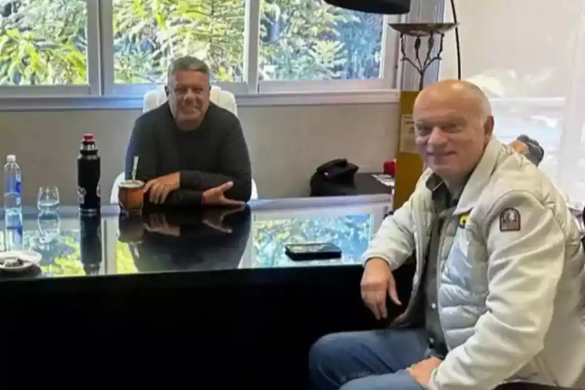 Two men sitting in an office across from a glass desk with windows in the background and objects like a water bottle, a thermos, and a mate on the table. Two men sitting in an office across from a glass desk with windows in the background and objects like a water bottle, a thermos, and a mate on the table.