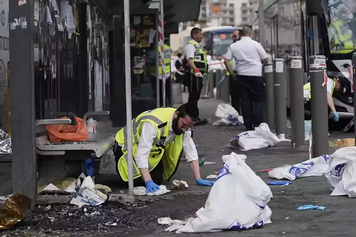A man wearing a yellow vest and blue gloves inspects the ground at a bus station surrounded by trash and bags while other people in similar vests watch the scene.