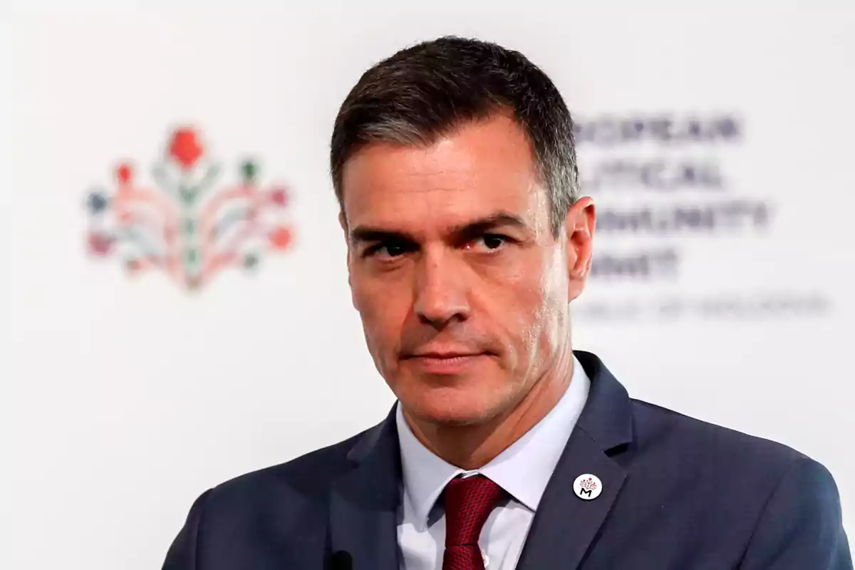 Short-haired, gray-haired man in a blue suit and red tie at an official event with a white background and a blurred colorful logo.