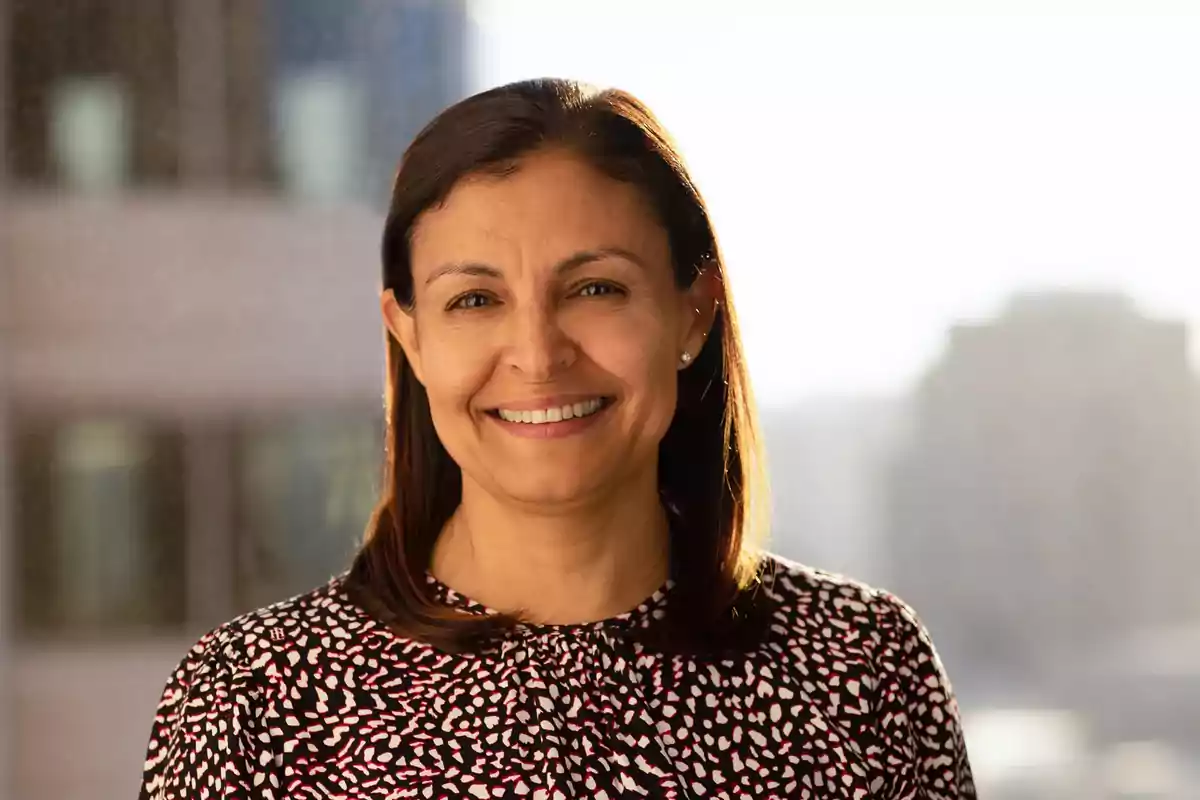 Mujer de cabello lacio y oscuro sonriendo, con blusa estampada, fondo de edificios desenfocados y luz natural Mujer de cabello lacio y oscuro sonriendo, con blusa estampada, fondo de edificios desenfocados y luz natural