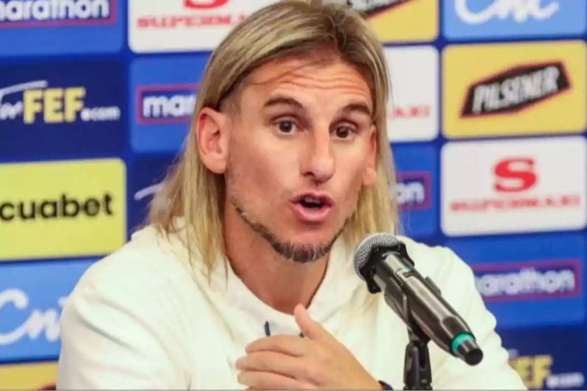 Hombre de cabello largo y rubio hablando frente a un micrófono en una rueda de prensa con fondo de logotipos deportivos