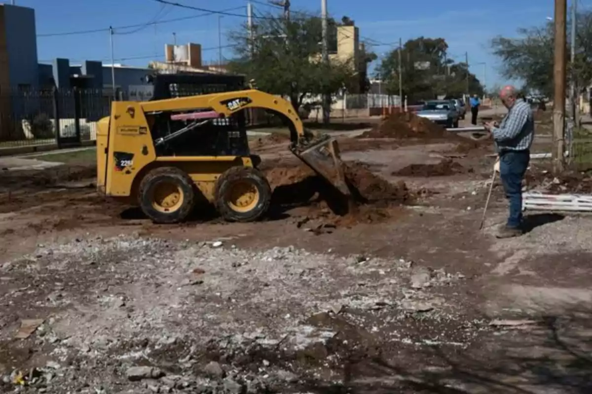 A skid steer loader moving dirt on a street under repair while a man watches and takes notes A skid steer loader moving dirt on a street under repair while a man watches and takes notes