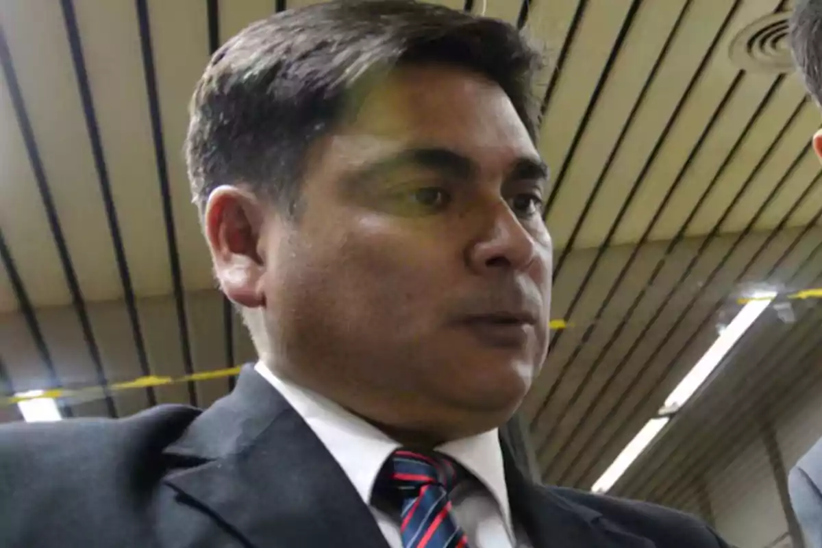Hombre de cabello corto y oscuro con traje y corbata en un entorno interior con techo de paneles Hombre de cabello corto y oscuro con traje y corbata en un entorno interior con techo de paneles