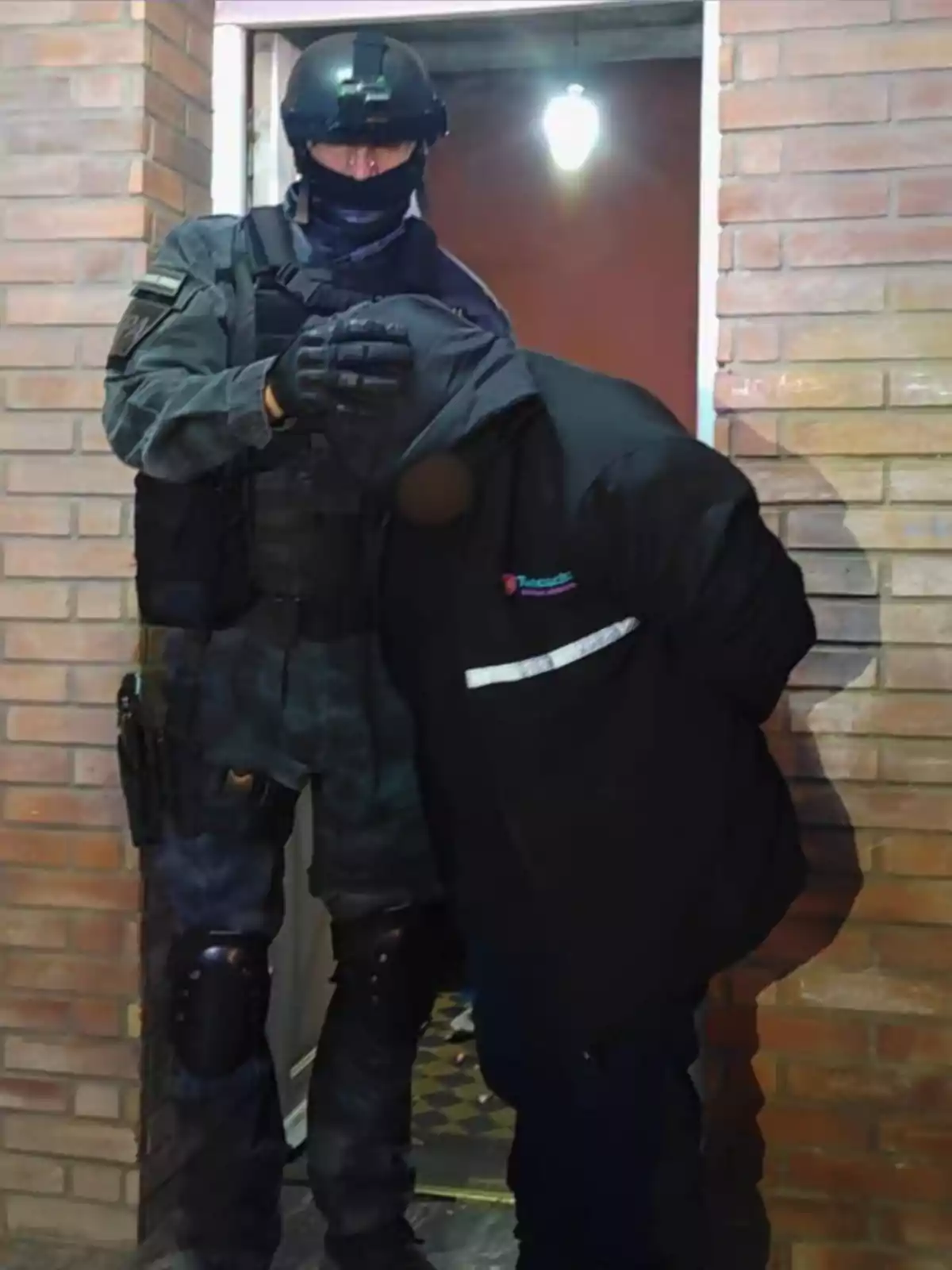 A police officer in tactical gear detains a hooded person at the entrance of a brick building. A police officer in tactical gear detains a hooded person at the entrance of a brick building.