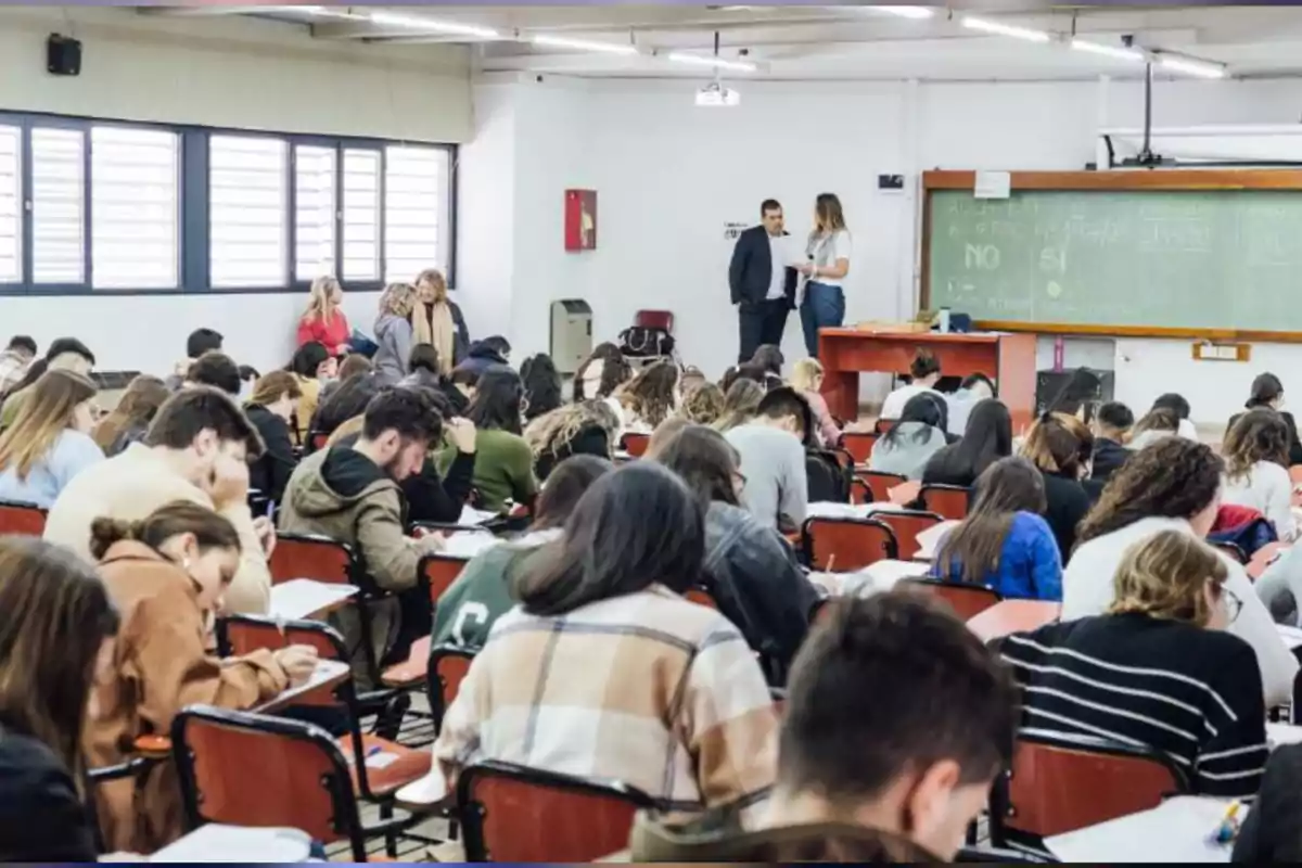 Aula llena de estudiantes sentados en pupitres tomando un examen mientras algunos profesores conversan cerca del pizarrón. Aula llena de estudiantes sentados en pupitres tomando un examen mientras algunos profesores conversan cerca del pizarrón.
