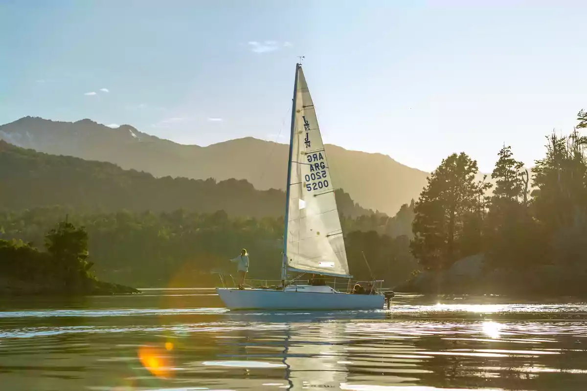 Velero navegando en un lago rodeado de montañas y árboles al atardecer Velero navegando en un lago rodeado de montañas y árboles al atardecer