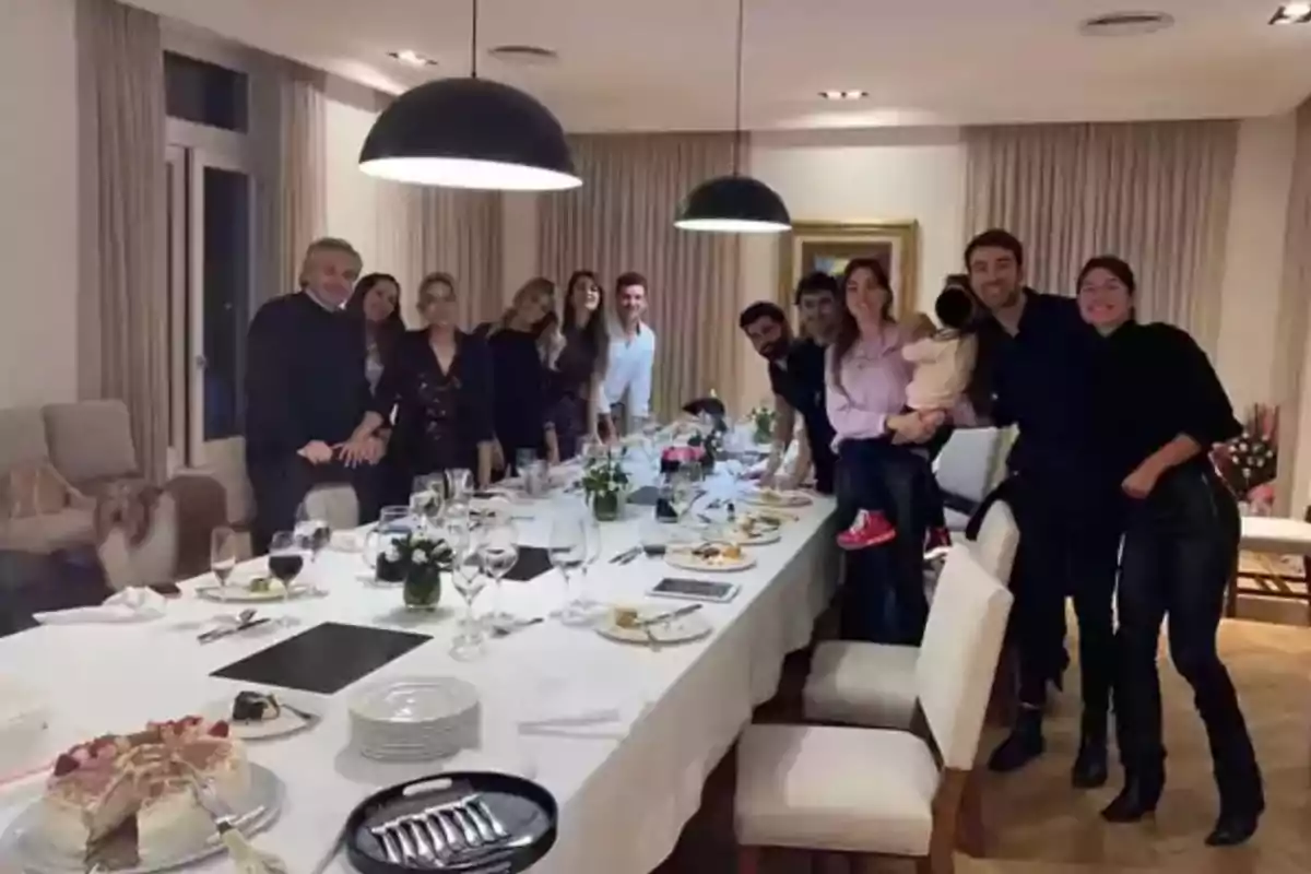 Un grupo de personas sonríe junto a una mesa larga con platos y copas después de una comida en un comedor elegante iluminado por lámparas colgantes Un grupo de personas sonríe junto a una mesa larga con platos y copas después de una comida en un comedor elegante iluminado por lámparas colgantes