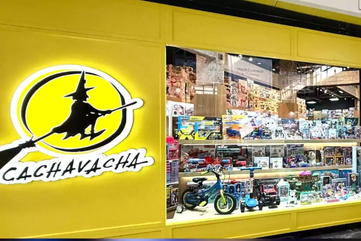 Fachada de una tienda de juguetes con un letrero amarillo que muestra la silueta de una bruja volando en escoba y vitrinas llenas de juguetes variados Fachada de una tienda de juguetes con un letrero amarillo que muestra la silueta de una bruja volando en escoba y vitrinas llenas de juguetes variados