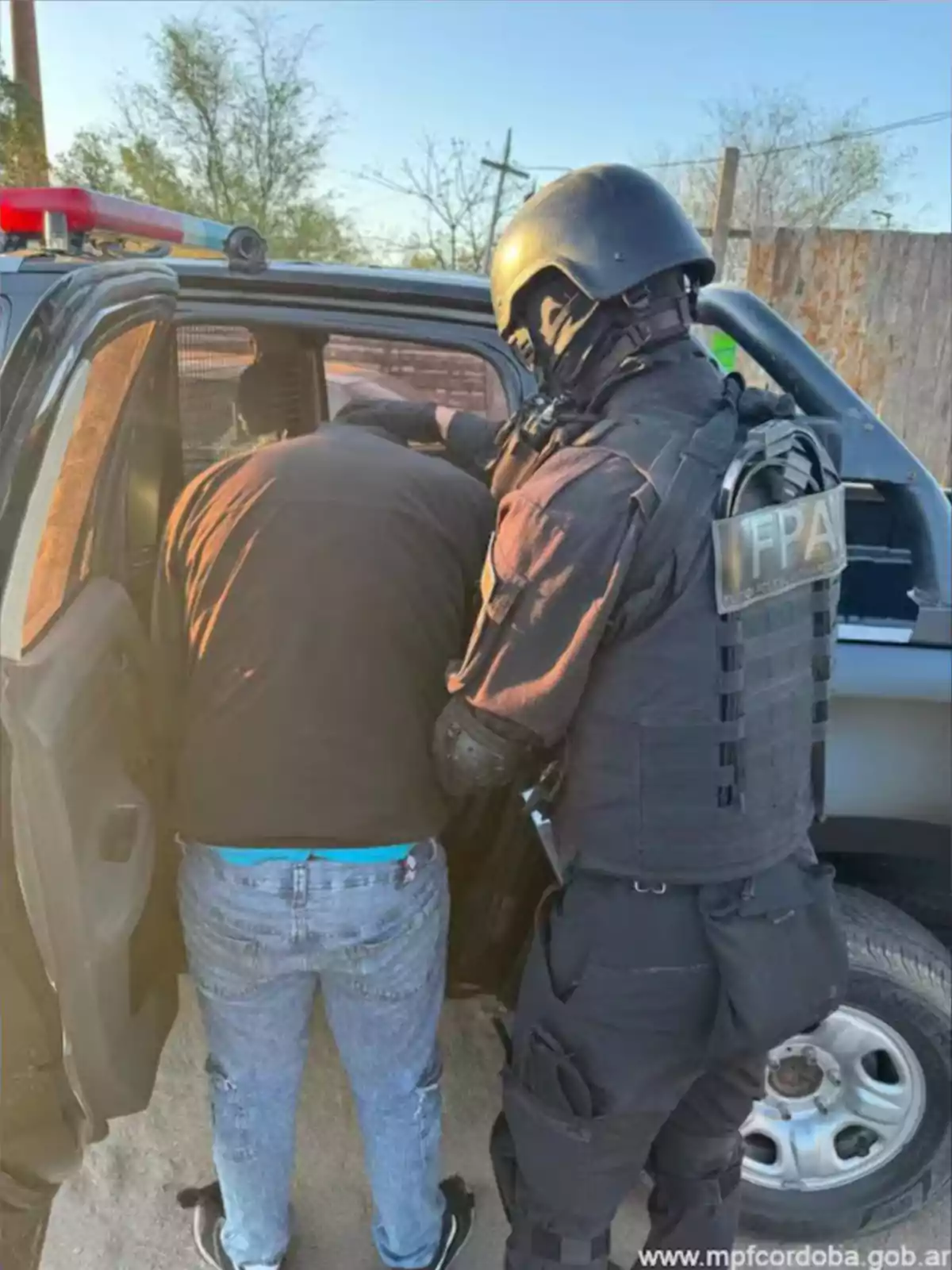 Un agente de seguridad con uniforme negro y casco escolta a una persona hacia el interior de una patrulla policial Un agente de seguridad con uniforme negro y casco escolta a una persona hacia el interior de una patrulla policial