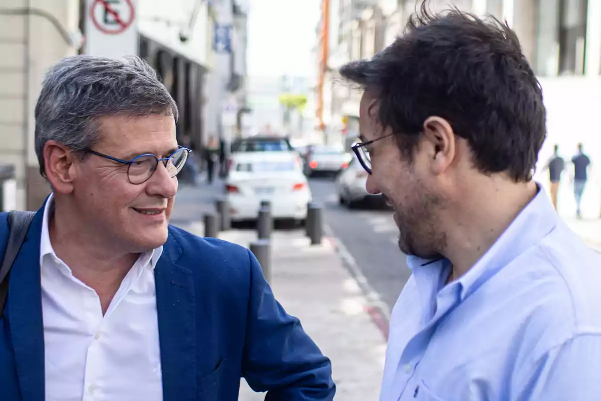 Two men with glasses talk and smile on an urban street during the day.