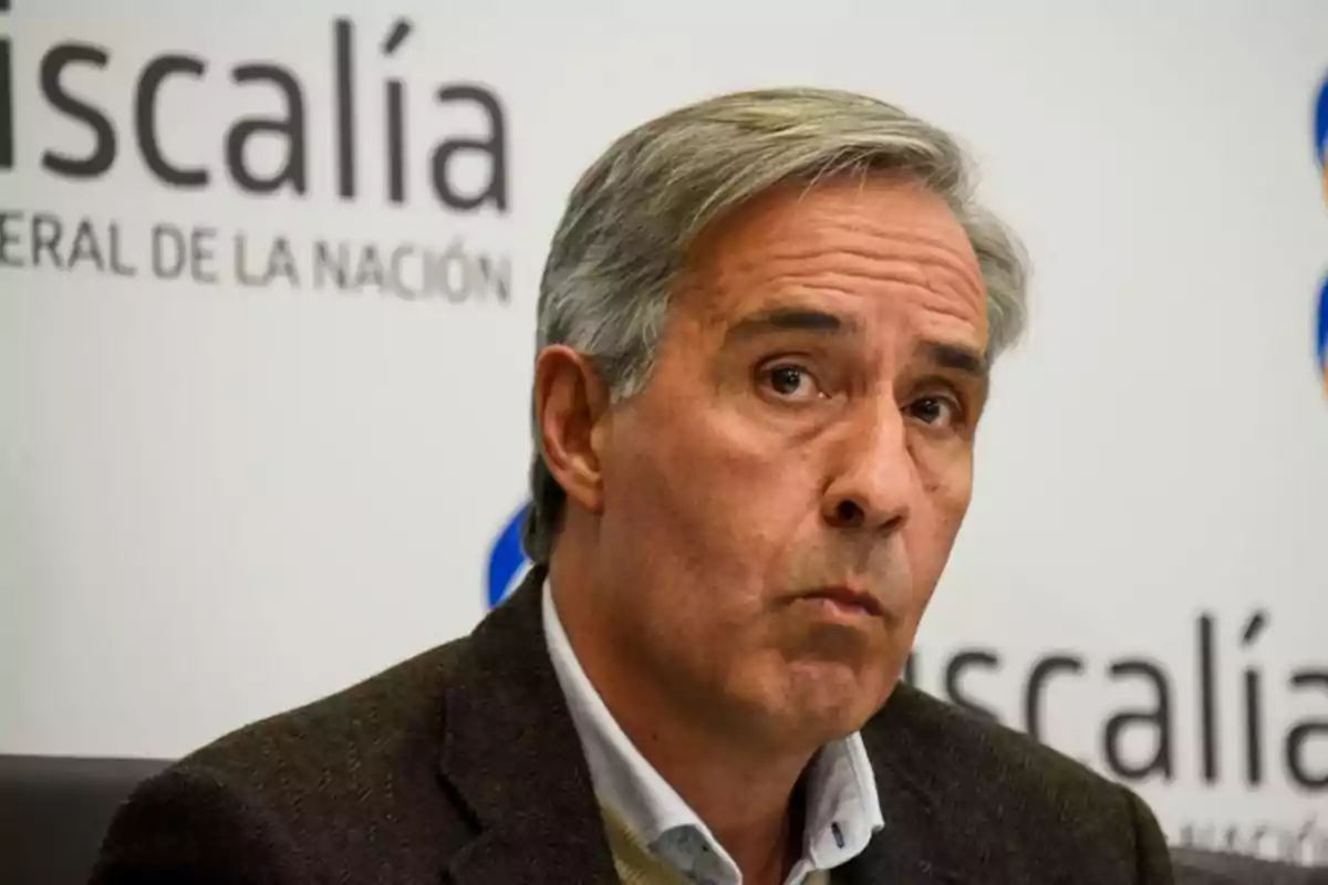 Hombre de cabello canoso con expresión seria sentado frente a un fondo con el texto fiscalía general de la nación Hombre de cabello canoso con expresión seria sentado frente a un fondo con el texto fiscalía general de la nación