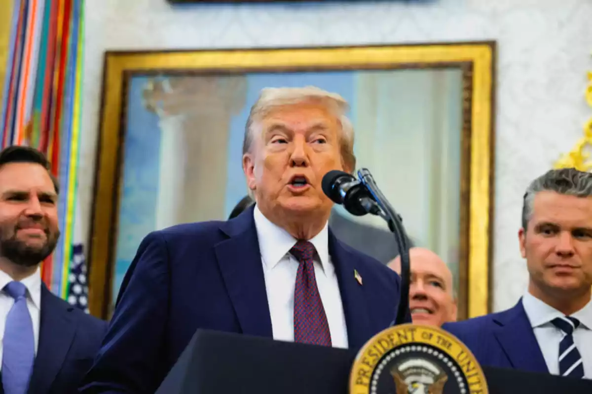 Un hombre de cabello rubio con traje azul habla frente a un micrófono en un podio presidencial acompañado de otras personas en un entorno formal con una pintura y banderas al fondo