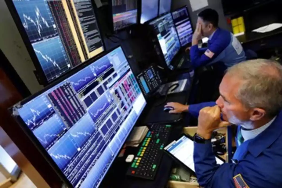 Two people in blue jackets observe several screens with financial charts in a trading office. Two people in blue jackets observe several screens with financial charts in a trading office.