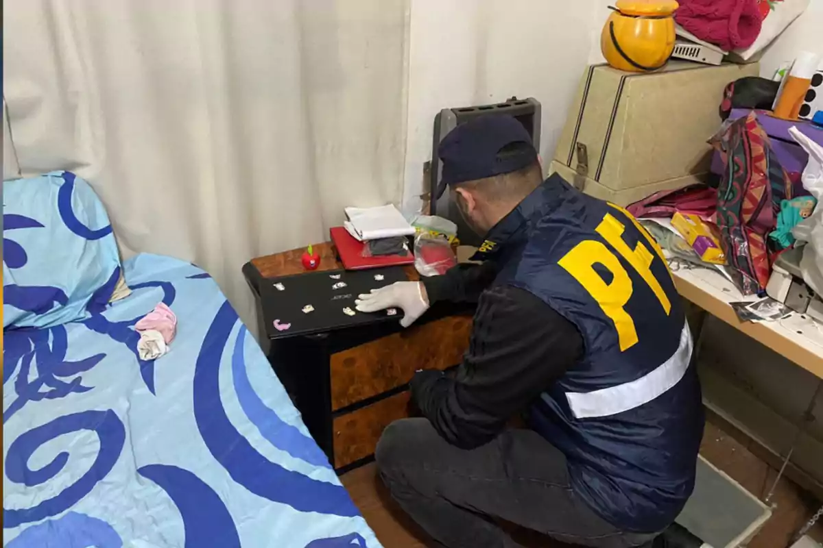 Agente con chaleco de la PFA revisando una computadora portátil en una habitación desordenada junto a una cama con sábanas azules
