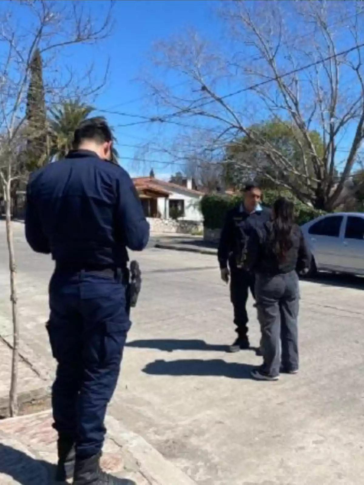 Three people, two of them in police uniforms, are talking on a residential street next to a white car and leafless trees. Three people, two of them in police uniforms, are talking on a residential street next to a white car and leafless trees.
