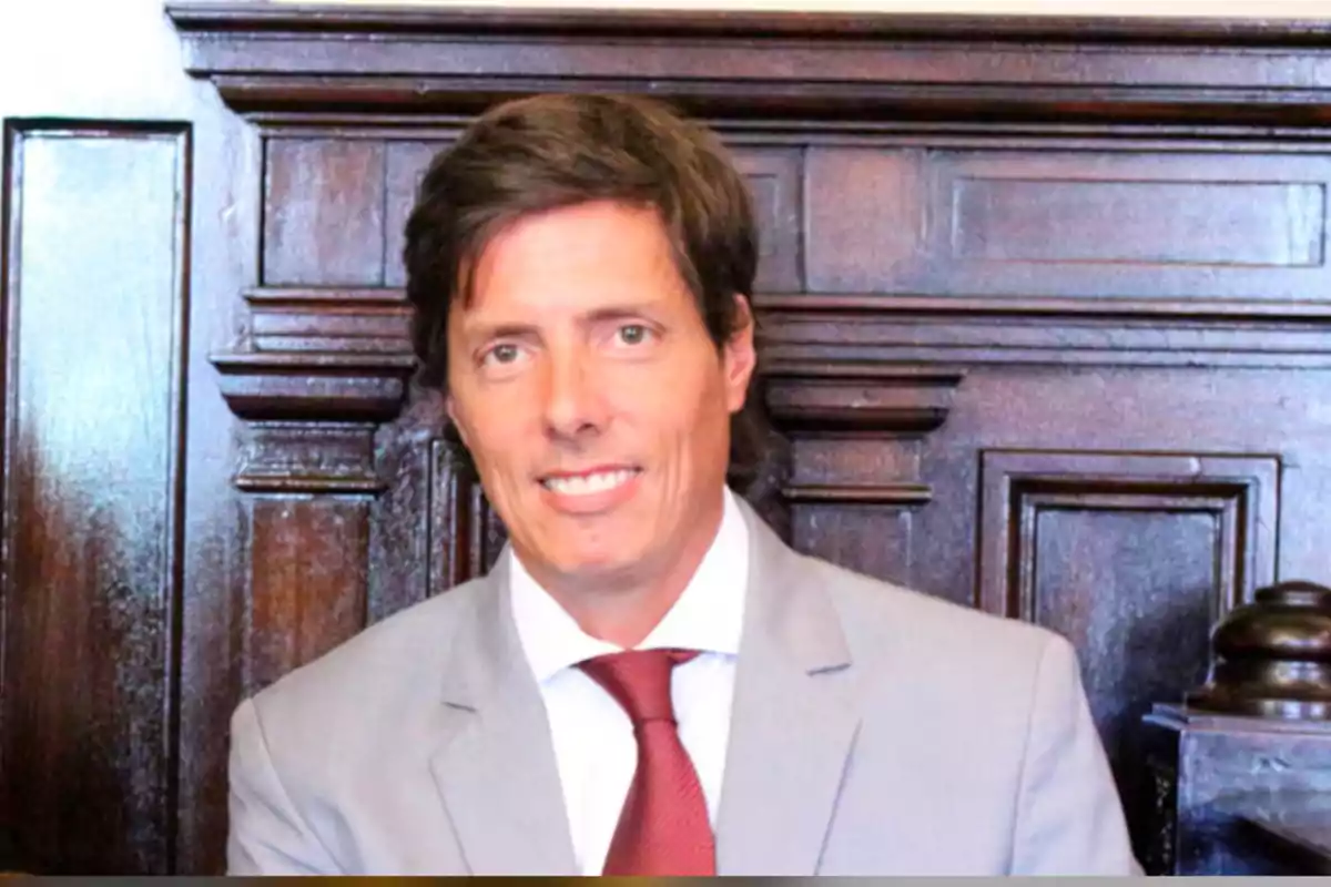 Hombre de cabello castaño con traje gris y corbata roja sonriendo frente a un fondo de madera oscura Hombre de cabello castaño con traje gris y corbata roja sonriendo frente a un fondo de madera oscura
