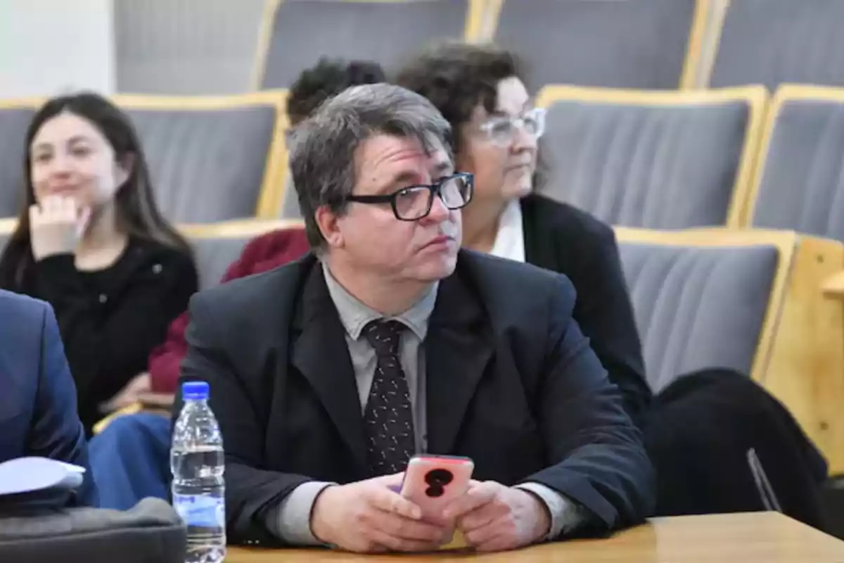 Hombre con gafas y traje sosteniendo un teléfono móvil sentado en una sala de conferencias con otras personas al fondo Hombre con gafas y traje sosteniendo un teléfono móvil sentado en una sala de conferencias con otras personas al fondo
