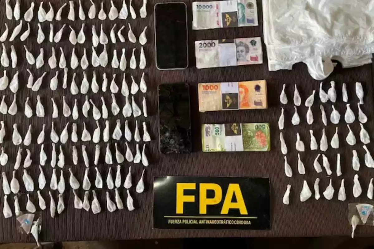 Paquetes pequeños envueltos en plástico blanco, dos teléfonos móviles, fajos de billetes, una prenda blanca y un cartel negro con letras amarillas que dice FPA sobre una mesa oscura Paquetes pequeños envueltos en plástico blanco, dos teléfonos móviles, fajos de billetes, una prenda blanca y un cartel negro con letras amarillas que dice FPA sobre una mesa oscura