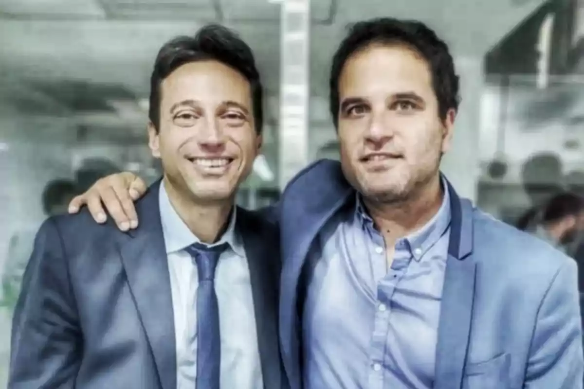 Two men dressed in suits smile and pose together in an office setting.
