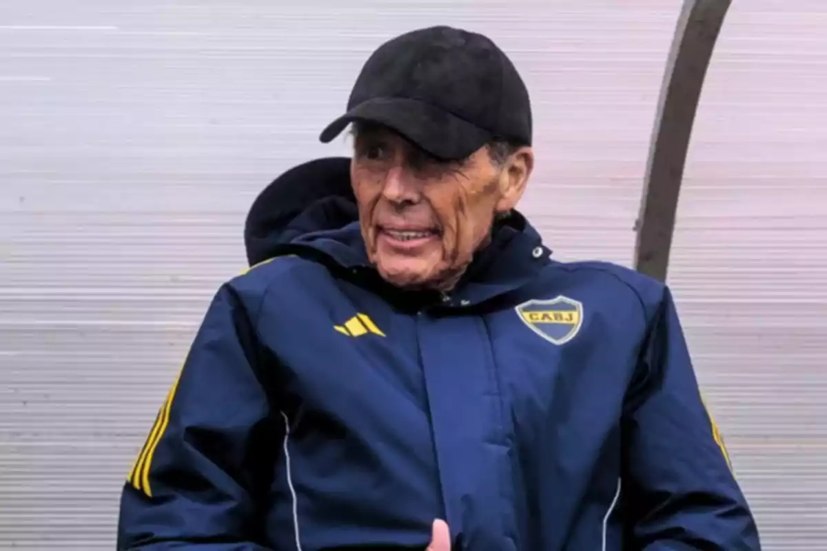 Older man wearing a black cap and a blue Boca Juniors jacket sitting on a substitute bench