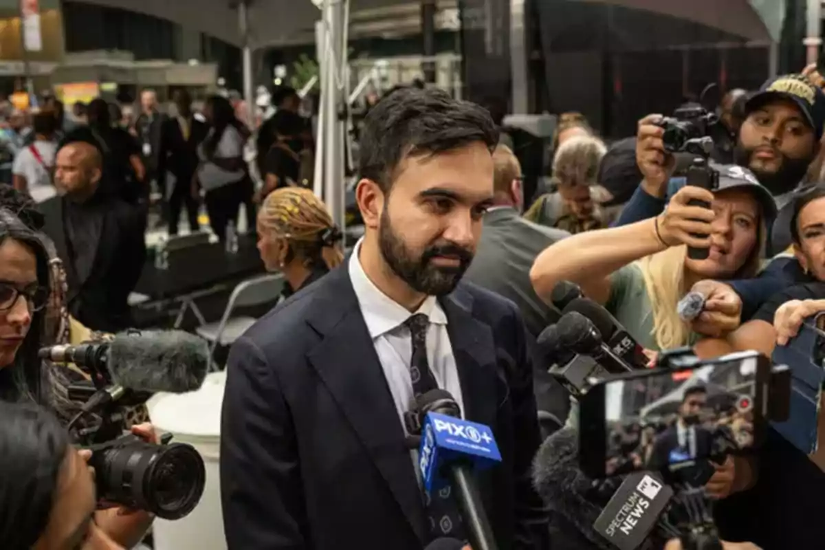 Hombre con barba y traje oscuro siendo entrevistado por varios periodistas y cámaras en un evento concurrido