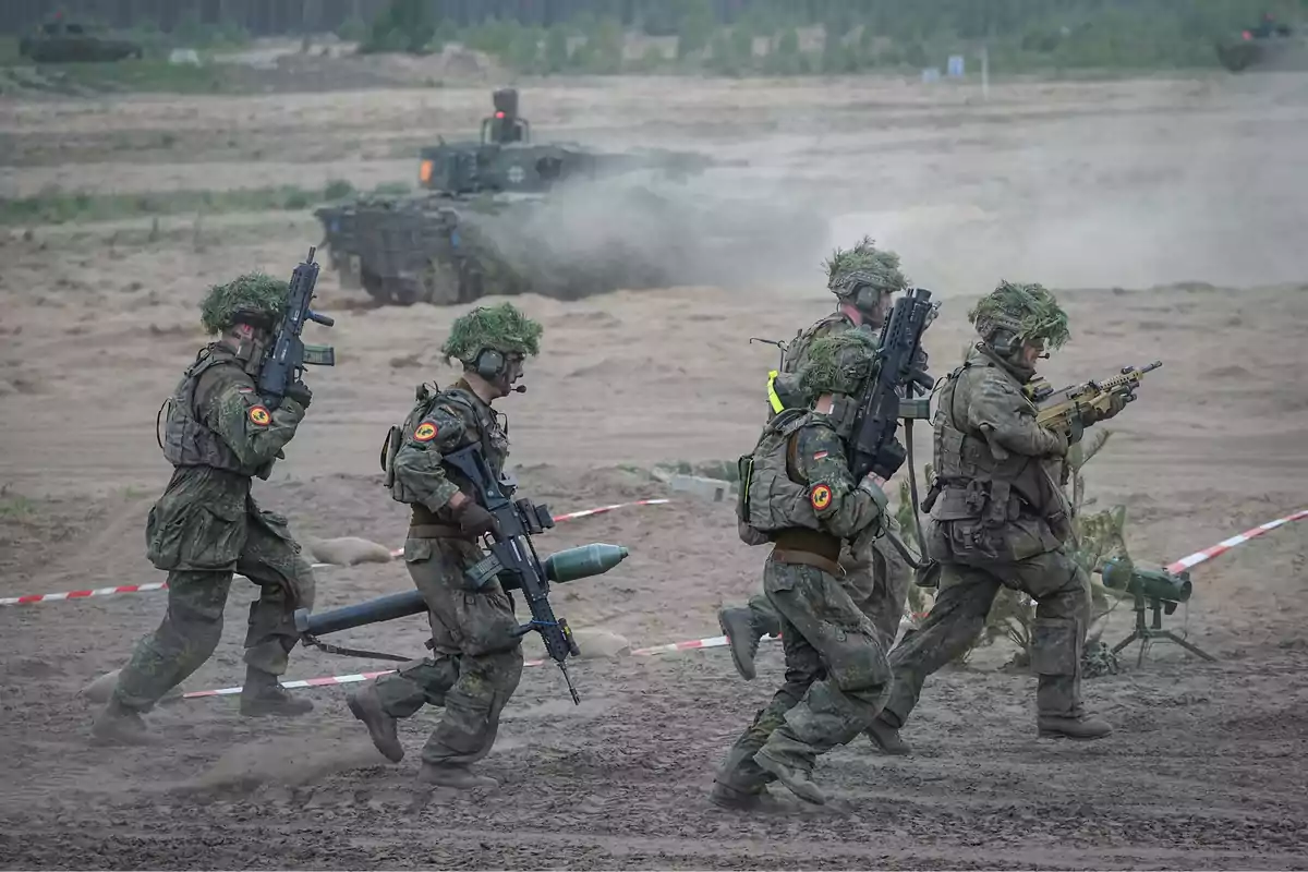 Soldados con uniforme de camuflaje y armas avanzan en formación sobre terreno arenoso mientras un vehículo blindado se observa al fondo entre polvo y vegetación dispersa Soldados con uniforme de camuflaje y armas avanzan en formación sobre terreno arenoso mientras un vehículo blindado se observa al fondo entre polvo y vegetación dispersa
