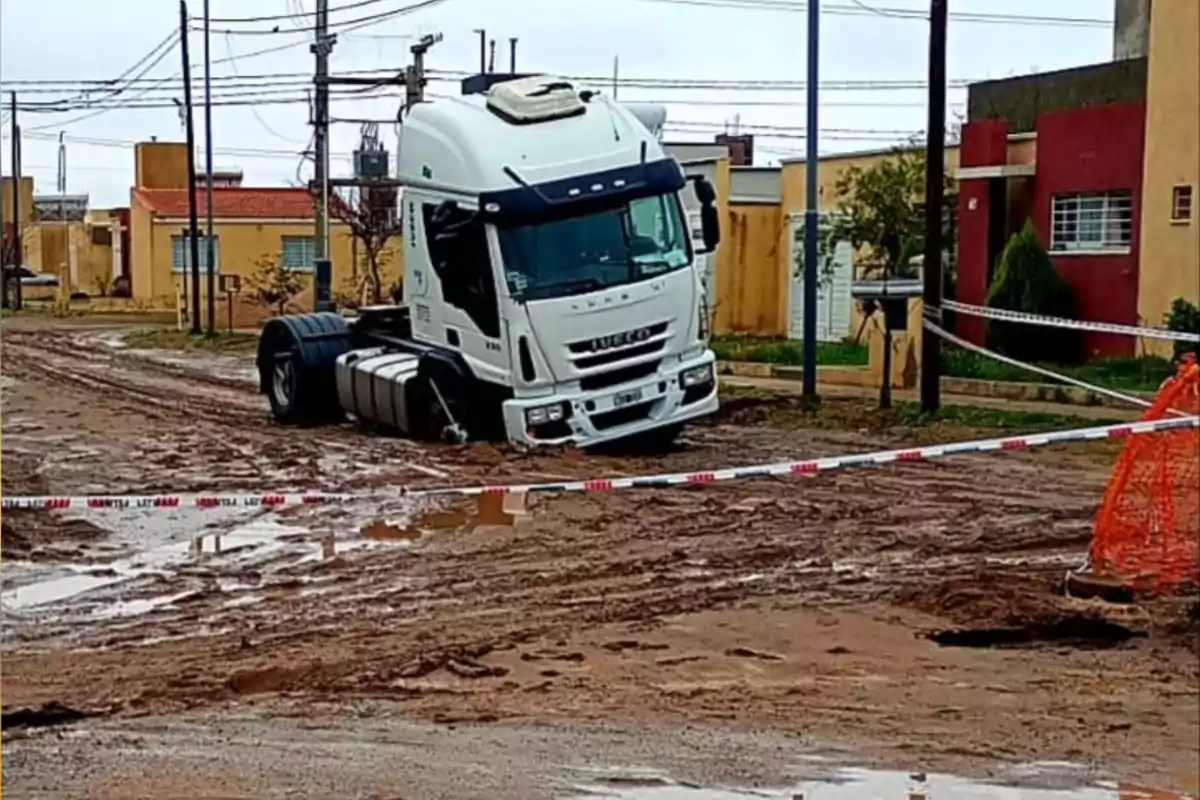 White truck stuck in the mud of an unpaved street in a residential area, surrounded by caution tape and puddles of water.