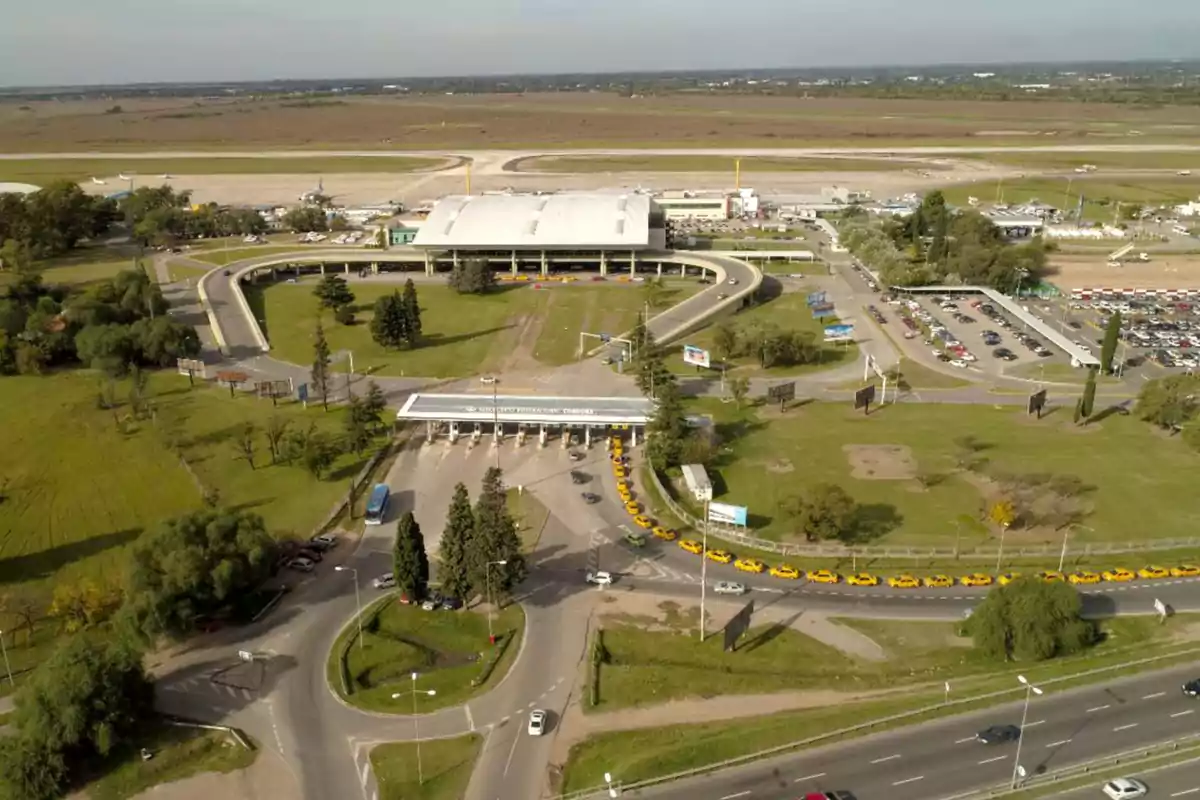 Aerial view of an airport with a main terminal, a line of yellow taxis waiting at the entrance, and green areas surrounding it. Aerial view of an airport with a main terminal, a line of yellow taxis waiting at the entrance, and green areas surrounding it.