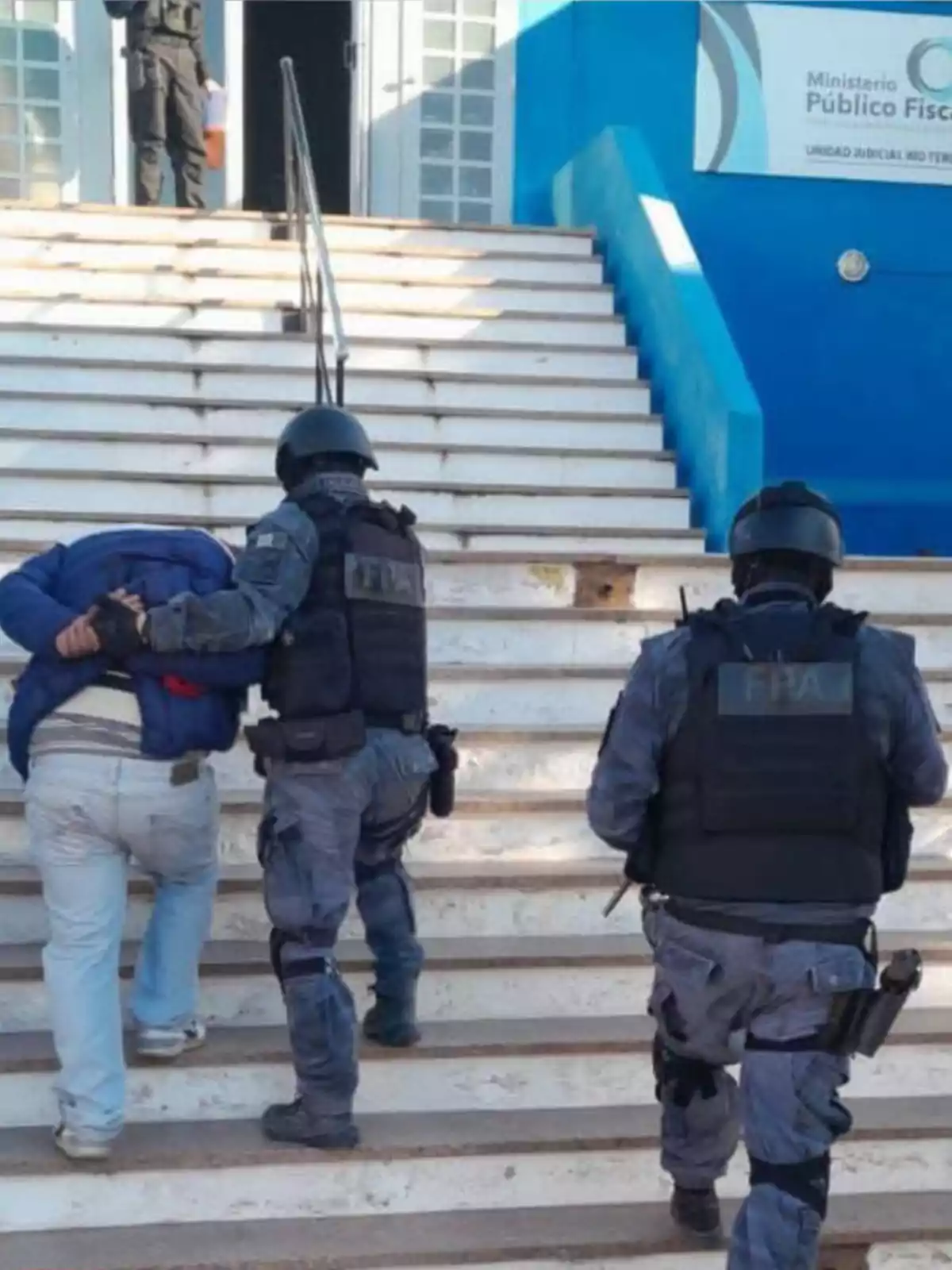 Police officers in tactical uniforms escort a handcuffed person up some stairs toward a blue government building. Police officers in tactical uniforms escort a handcuffed person up some stairs toward a blue government building.
