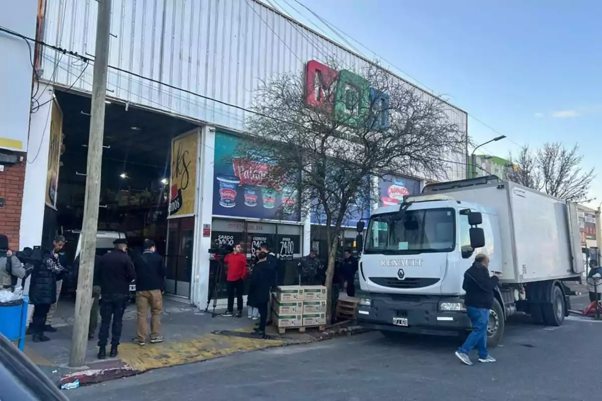 Fachada de un supermercado mayorista con un camión de reparto estacionado en la entrada y varias personas reunidas en la acera
