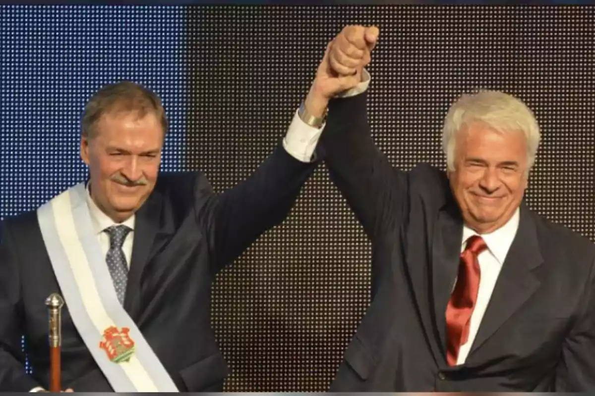 Two men in suits smile and raise their clasped hands in a gesture of celebration. Two men in suits smile and raise their clasped hands in a gesture of celebration.