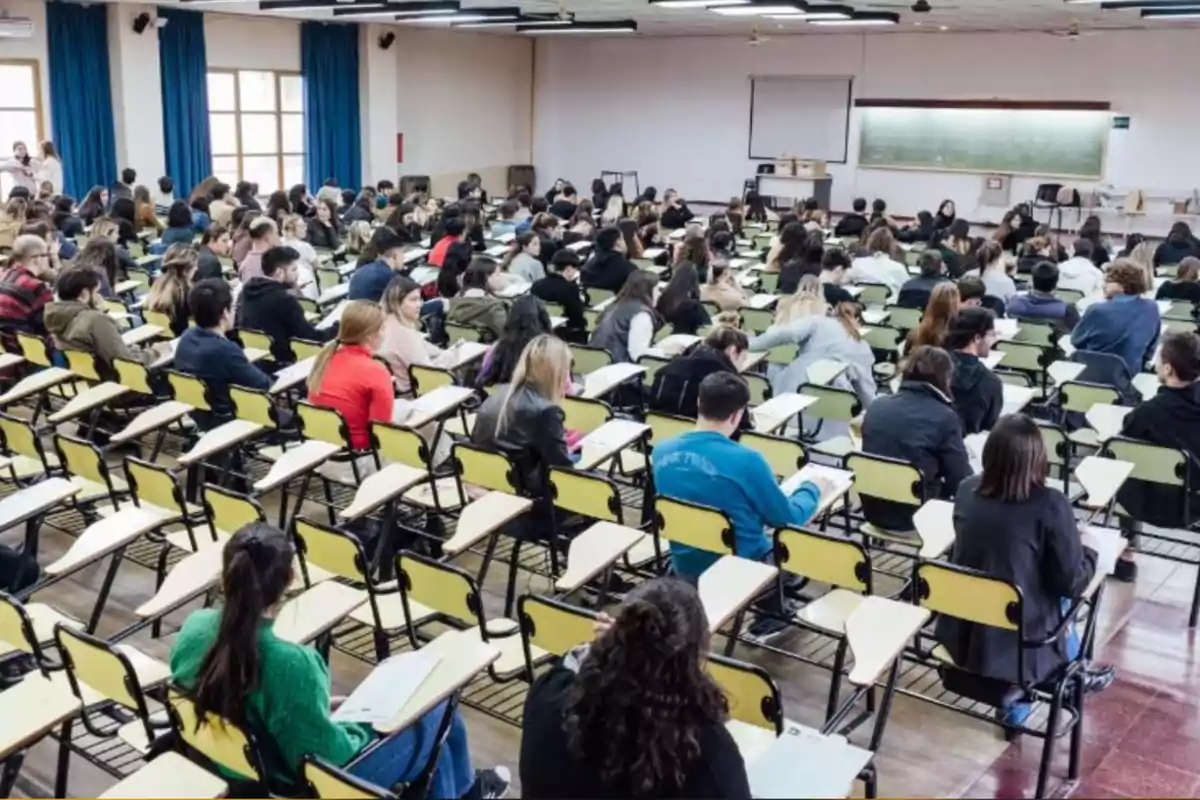Aula universitaria llena de estudiantes sentados en pupitres tomando apuntes frente a un pizarrón y una pantalla de proyección