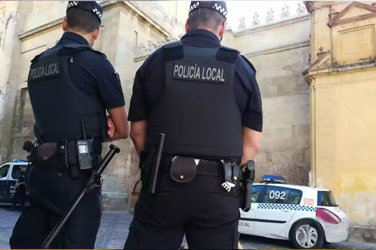 Two local police officers with their backs turned on a street next to a patrol car Two local police officers with their backs turned on a street next to a patrol car
